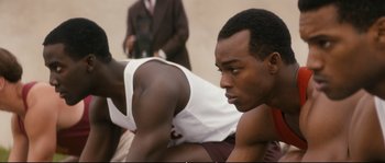 Movie still from “Race” (2016), directed by Stephen Hopkins – A man sitting on the ground with another man standing next to him; Close Up shot, Over the shoulder angle