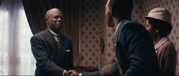 Movie still from “Race” (2016), directed by Stephen Hopkins – Two men in suits shaking hands in front of a wall; Medium shot, Over the shoulder angle