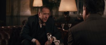 Movie still from “Race” (2016), directed by Stephen Hopkins – An older man sitting at a table in front of a lamp; Medium shot, Over the shoulder angle