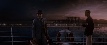 Movie still from “Race” (2016), directed by Stephen Hopkins – A man in a suit and hat standing on a boat in the water; Wide shot, Low angle