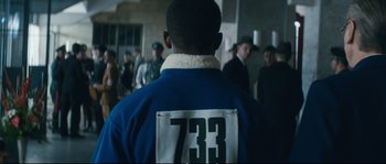Movie still from “Race” (2016), directed by Stephen Hopkins – A man in a blue jacket is standing in front of a group of people; Medium shot, Over the shoulder angle