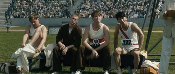 Movie still from “Race” (2016), directed by Stephen Hopkins – A group of young men sitting on top of a grass covered field; Medium shot, High angle