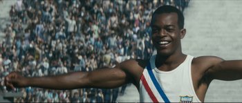 Movie still from “Race” (2016), directed by Stephen Hopkins – A person smiling in front of a crowd; Medium shot, Low angle