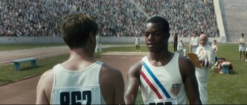 Movie still from “Race” (2016), directed by Stephen Hopkins – Two men standing next to each other in front of a crowd; Medium shot, Over the shoulder angle