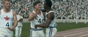 Movie still from “Race” (2016), directed by Stephen Hopkins – A couple of men standing next to each other on top of a field; Medium shot, Over the shoulder angle