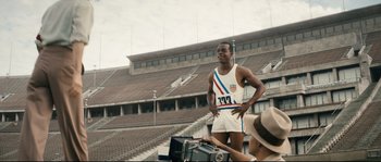 Movie still from “Race” (2016), directed by Stephen Hopkins – A man standing on a field with a camera in front of him; Medium shot, Low angle