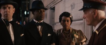 Movie still from “Race” (2016), directed by Stephen Hopkins – A man and a woman standing next to each other in front of a building; Medium shot, Low angle