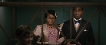Movie still from “Race” (2016), directed by Stephen Hopkins – A woman in a pink dress standing next to a man in a suit; Medium shot, Over the shoulder angle