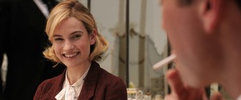 Movie still from “Rebecca” (2020), directed by Ben Wheatley – A young woman smiles as she sits in front of a mirror; Close Up shot, Over the shoulder angle