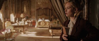 Movie still from “Rebecca” (2020), directed by Ben Wheatley – A woman standing in a room holding a book in her hands; Medium shot, Over the shoulder angle