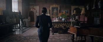 Movie still from “Rebecca” (2020), directed by Ben Wheatley – A woman in a suit standing in front of a painting in a living room; Wide shot, Over the shoulder angle
