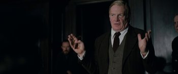 Movie still from “Rebecca” (2020), directed by Ben Wheatley – A man in a suit and tie holding his hand out; Medium shot, Over the shoulder angle