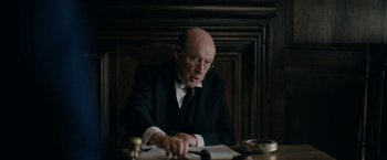 Movie still from “Rebecca” (2020), directed by Ben Wheatley – An older man sitting at a table writing on a piece of paper; Medium shot, Over the shoulder angle
