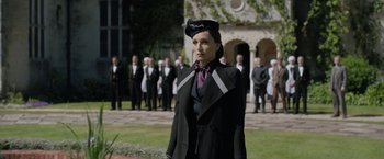 Movie still from “Rebecca” (2020), directed by Ben Wheatley – A woman wearing a hat and a suit stands in front of a group of men; Medium shot, Low angle