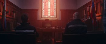 Movie still from “Red Notice” (2021), directed by Rawson Marshall Thurber – Two people sitting at a desk in front of a window; Wide shot, Over the shoulder angle