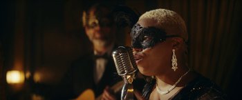 Movie still from “Red Notice” (2021), directed by Rawson Marshall Thurber – A woman wearing a black mask and a black mask is singing into a microphone while a man wearing a black mask is playing a guitar; Close Up shot, Low angle