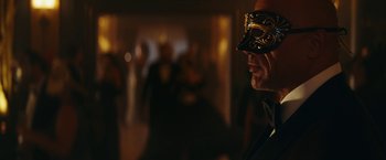Movie still from “Red Notice” (2021), directed by Rawson Marshall Thurber – A man wearing a mask in front of a group of people; Close Up shot, Over the shoulder angle