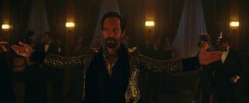 Movie still from “Red Notice” (2021), directed by Rawson Marshall Thurber – A man with a beard and a suit and tie in front of a group of people; Medium shot, Over the shoulder angle