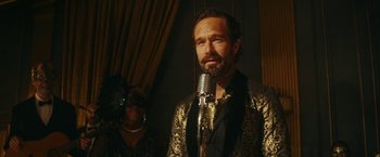 Movie still from “Red Notice” (2021), directed by Rawson Marshall Thurber – A man in a gold jacket is speaking into a microphone; Close Up shot, Over the shoulder angle