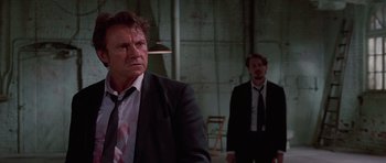 Movie still from “Reservoir Dogs” (1992), directed by Quentin Tarantino – A man in a suit and tie standing next to another man in a suit and tie; Medium shot, Low angle