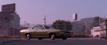 Movie still from “Reservoir Dogs” (1992), directed by Quentin Tarantino – An old car driving down a street with a fence in the background; Wide shot, Low angle