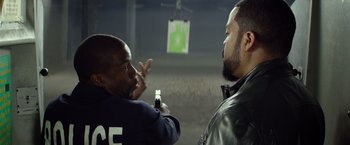 Movie still from “Ride Along” (2014), directed by Tim Story – A man holding a gun pointed at another man in front of a shooting target; Medium shot, Over the shoulder angle