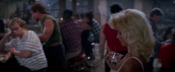 Movie still from “Road House” (1989), directed by Rowdy Herrington – A woman holding a drink in her hand while standing next to a crowd; Medium shot, Over the shoulder angle