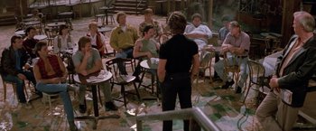 Movie still from “Road House” (1989), directed by Rowdy Herrington – A group of people sitting in a circle with a woman standing in front of them; Wide shot, High angle