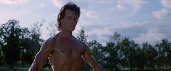 Movie still from “Road House” (1989), directed by Rowdy Herrington – A shirtless man standing in a field with trees in the background; Medium shot, Low angle