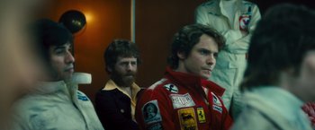 Movie still from “Rush” (2013), directed by Ron Howard – A man with a beard is standing next to a man with a red jacket; Medium shot, Over the shoulder angle