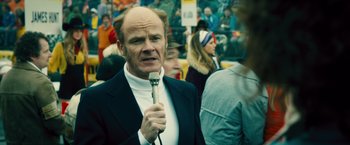 Movie still from “Rush” (2013), directed by Ron Howard – A man in a suit holding a microphone in front of a crowd of onlookers; Close Up shot, Over the shoulder angle