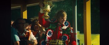Movie still from “Rush” (2013), directed by Ron Howard – A man in a red jacket holding a trophy; Medium shot, Low angle