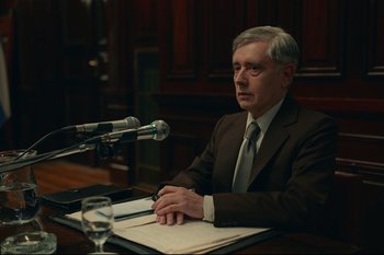 Movie still from “Argentina, 1985” (2022), directed by Santiago Mitre – A man sitting at a table in front of microphones; Medium shot, Over the shoulder angle