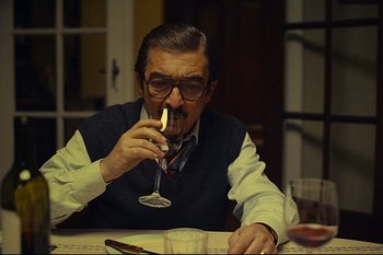 Movie still from “Argentina, 1985” (2022), directed by Santiago Mitre – An older man sitting at a table drinking a glass of wine; Medium shot, Over the shoulder angle