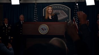 Movie still from “Salvation” (2017), created by Elizabeth Kruger – A woman standing at a podium in front of an american flag; Medium shot, Over the shoulder angle