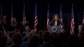 Movie still from “Salvation” (2017), created by Elizabeth Kruger – A woman standing at a podium in front of a group of people; Medium shot, Over the shoulder angle