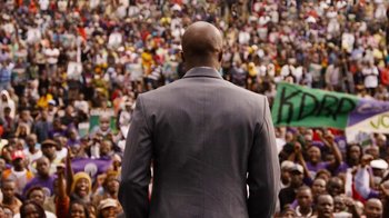 Movie still from “Sense8” (2015), directed by Tom Tykwer – A man in a suit standing in front of a crowd of onlookers; Medium shot, Over the shoulder angle