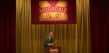 Movie still from “Sex Education” (2019), created by Laurie Nunn – A man standing at a podium in front of a banner that reads " moordale secondary "; Extreme Wide shot, High angle