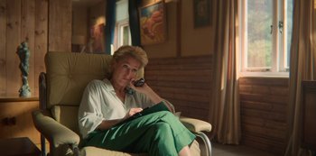 Movie still from “Sex Education” (2019), created by Laurie Nunn – A woman sitting in a chair holding a book and talking on the phone; Medium shot, Low angle