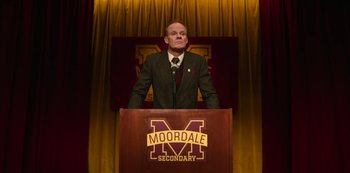 Movie still from “Sex Education” (2019), created by Laurie Nunn – A man standing at a podium in front of a moordale sign; Medium shot, High angle