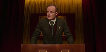 Movie still from “Sex Education” (2019), created by Laurie Nunn – A man standing at a podium in front of a microphone; Medium shot, Low angle