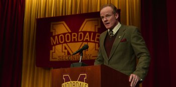 Movie still from “Sex Education” (2019), created by Laurie Nunn – A man standing at a podium with a microphone in front of him; Medium shot, High angle