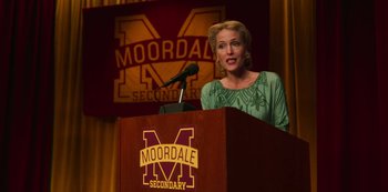 Movie still from “Sex Education” (2019), created by Laurie Nunn – A woman standing at a podium with a microphone; Medium shot, High angle
