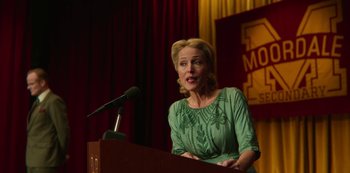 Movie still from “Sex Education” (2019), created by Laurie Nunn – A woman in a green dress is speaking at a podium; Medium shot, Over the shoulder angle