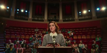 Movie still from “Sex Education” (2019), created by Laurie Nunn – A woman is speaking at a podium in front of an audience; Medium shot, Low angle