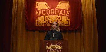 Movie still from “Sex Education” (2019), created by Laurie Nunn – A woman standing at a lectern in front of a moordale banner; Medium shot, High angle