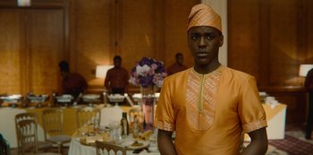 Movie still from “Sex Education” (2019), created by Laurie Nunn – A man in an orange and yellow outfit standing in front of a table; Medium shot, Low angle