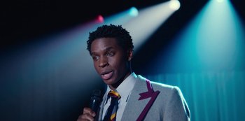 Movie still from “Sex Education” (2019), created by Laurie Nunn – A man in a suit holding a microphone in front of lights; Close Up shot, Low angle