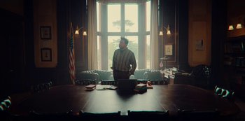 Movie still from “Sex Education” (2019), created by Laurie Nunn – A man sitting at a table in front of an american flag; Wide shot, Low angle