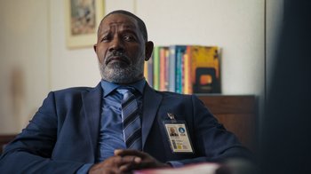 Movie still from “Sniper: Rogue Mission” (2022), directed by Oliver Thompson – A man in a suit and tie sitting in front of a bookshelf; Close Up shot, Over the shoulder angle